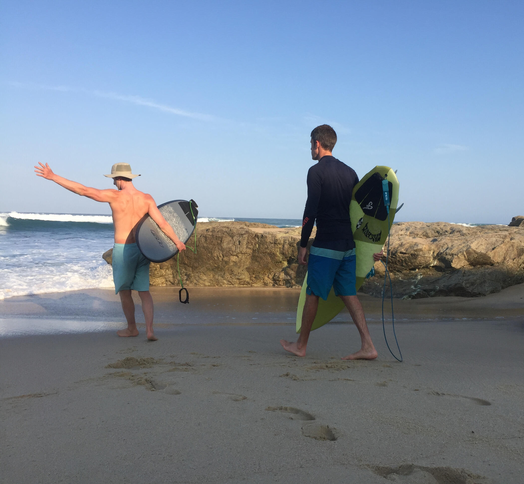 Two surfers walking down a rocky beach with boards, friendly, relaxed energy